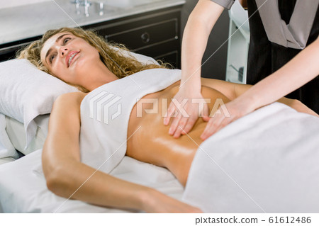 Beautiful blond curly woman smiling, while lying on the bed and having stomach massage. Body care, medical spa center. Beautiful blond curly woman smiling, while lying on the bed and having stomach massage. Body care, medical spa center. 61612486