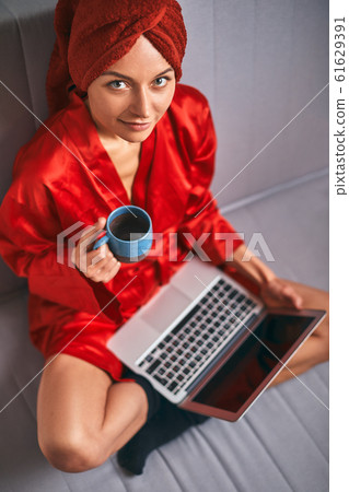 Young woman in red bathrobe and red towel on her head is standing in kitchen near table, drinking coffee and using laptop. Morning, girl after shower drinks tea and works on computer.Freelancer works Young woman in red bathrobe and red towel on her head is standing in kitchen near table, drinking coffee and using laptop. Morning, girl after shower drinks tea and works on computer.Freelancer works 61629391