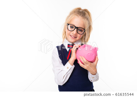 Pretty schoolgirl with blonde hair in school suit smiles and holds pink pig money box with money Pretty schoolgirl with blonde hair in school suit smiles and holds pink pig money box with money 61635544