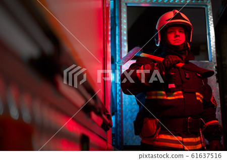 Firefighter male with hammer on his shoulder standing near fire engine. Firefighter male with hammer on his shoulder standing near fire engine. 61637516