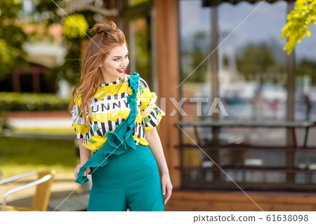 Woman close up stands against the background of the city cafeteria in a bright yellow tank top and turquoise overalls 61638098