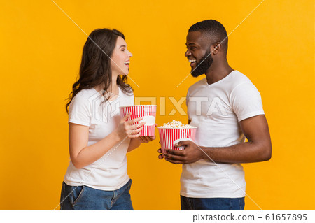 Cheerful Interracial Couple Holding Popcorn Buckets And Laughing Over Yellow Background 61657895