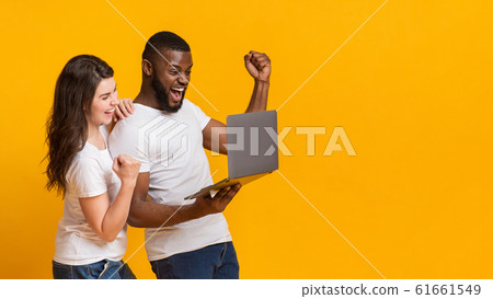 Happy interracial spouses celebrating win with laptop, raising fists in excitement 61661549
