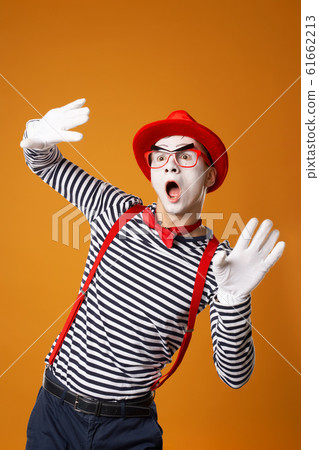 Young mime male in white gloves and red hat looking at side on orange background Young mime male in white gloves and red hat looking at side on orange background 61662213