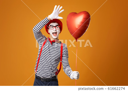 Happy mime male in red hat and vest holds heart ball in his hands on empty orange background 61662274