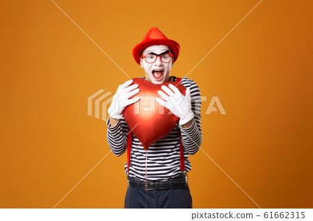 Sad clown mime in red hat and vest holds heart ball in his hands on orange background 61662315