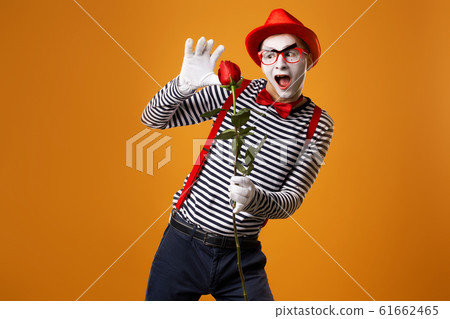 Funny mime with white face in red hat and striped t-shirt holding rose isolated on empty orange background Funny mime with white face in red hat and striped t-shirt holding rose isolated on empty orange background 61662465