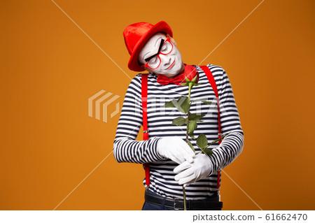 Smilling mime man with white face in red hat and striped t-shirt holding rose isolated on empty orange background 61662470