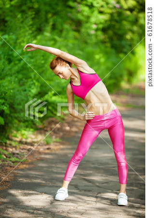 Training In The Fresh Air in The Park. The Sports Girl in a Pink Undershirt, Pink Leggings and White Neakers Against the Background of Green Foliage. The Woman Does Warm-up and Charging 61678518