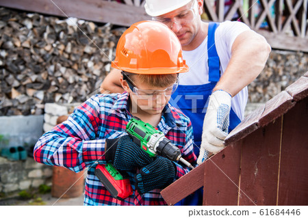 Father and son are building a doghouse together. Father is teaching his son to use the electric screwdriver. Father and son are building a doghouse together. Father is teaching his son to use the electric screwdriver. 61684446