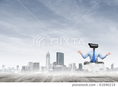 Camera headed man sitting in lotus pose on cloud against modern cityscape 61690219