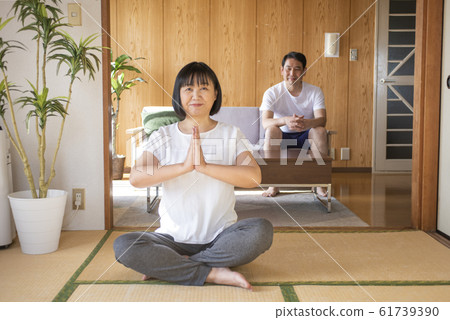 Middle-aged woman doing stretching yoga at home and her husband watching it Middle-aged woman doing stretching yoga at home and her husband watching it 61739390