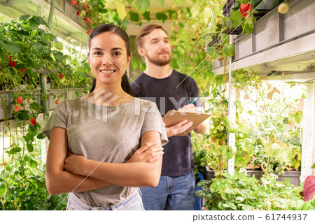 Young cheerful female farmer looking at you on background of her colleague 61744937