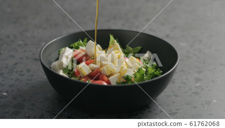 add olive oil to salad with mozzarella, cherry tomatoes and frisee leaves in black bowl on terrazzo surface 61762068