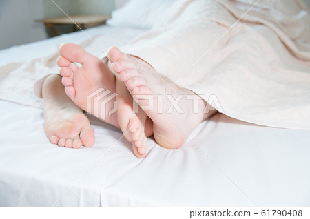 Close-up of the feet of a young couple sticking out from under the covers in the bedroom. Bare feet caress each other engaged in grooming. 61790408