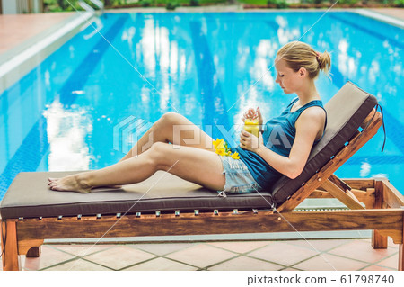 Young woman is holding a smoothie of mango on the background of the pool. Fruit smoothie - healthy eating concept. Close up of detox smoothie with mango Young woman is holding a smoothie of mango on the background of the pool. Fruit smoothie - healthy eating concept. Close up of detox smoothie with mango 61798740