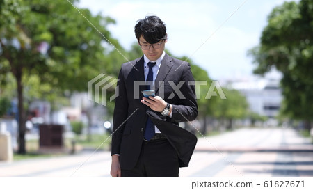A Japanese man in his 20s wearing a suit wearing glasses looking at a smartphone on the way to work on a sunny morning in front of the station A Japanese man in his 20s wearing a suit wearing glasses looking at a smartphone on the way to work on a sunny morning in front of the station 61827671