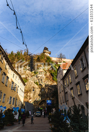 Graz, Styria - Austria - 24.12.2019: City of Graz street and stairway to Clock tower Uhrturm view, Schlossberg clock in background at hill, tourists walking restaurants. Graz, Styria - Austria - 24.12.2019: City of Graz street and stairway to Clock tower Uhrturm view, Schlossberg clock in background at hill, tourists walking restaurants. 61833144