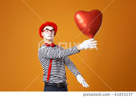 Serious clown mime in red hat and vest holds heart ball in his hands 61833237