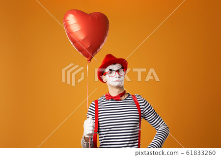Mime in red hat and vest holds heart ball in his hands on orange background Mime in red hat and vest holds heart ball in his hands on orange background 61833260