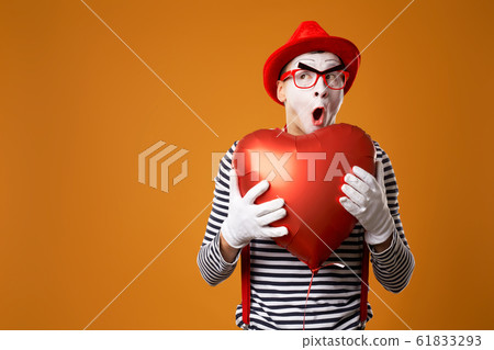 Close up surprised mime in red hat and vest holds heart ball in his hands 61833293