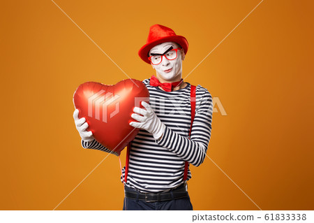 Happy clown mime in red hat and vest holds heart ball in his hands on orange background 61833338