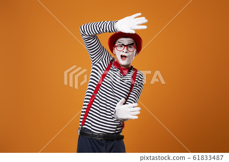 Mime in vest and red hat with invisible glass in hands on orange empty background 61833487
