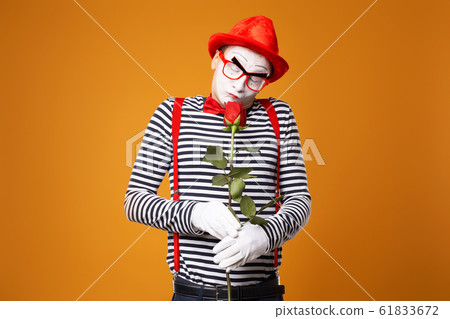 Pensive mime man with white face in red hat and striped t-shirt holding rose isolated on empty orange background 61833672