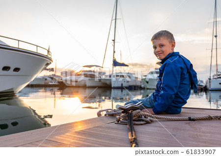 A boy with a smile in a blue jacket sits on a pier during sunset 61833907