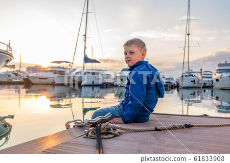 A boy in a blue jacket sits on a pier with yachts during sunset A boy in a blue jacket sits on a pier with yachts during sunset 61833908