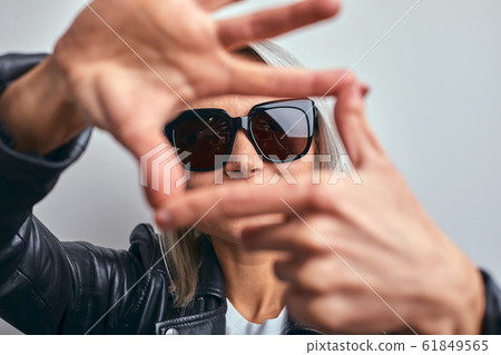 Beautiful woman in glasses, in a black leather jacket on a gray background, making a frame using hands with palms and fingers, camera perspective. Beautiful woman in glasses, in a black leather jacket on a gray background, making a frame using hands with palms and fingers, camera perspective. 61849565