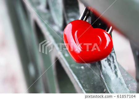 Red Valentine's Day Heart shape wedding lock hangs on metal bridge of lovers on blurred background. Red Valentine's Day Heart shape wedding lock hangs on metal bridge of lovers on blurred background. 61857104