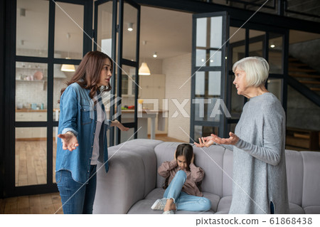 Mom and grandmother talking seriously, daughter covering her ears. Mom and grandmother talking seriously, daughter covering her ears. 61868438