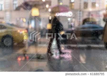 Blurry reflection silhouettes of the people in walking on a rain under umbrellas and bokeh city lights, night. View through glass window with rain drops Blurry reflection silhouettes of the people in walking on a rain under umbrellas and bokeh city lights, night. View through glass window with rain drops 61880795