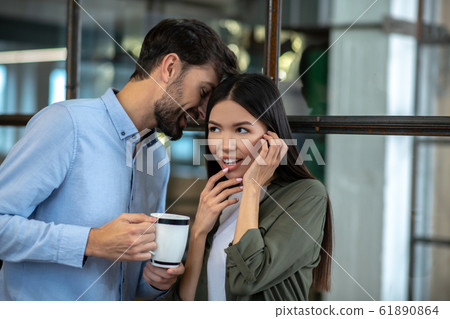 Tall young man having coffee and whispering something in the ear to his colleague 61890864