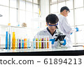 Science, Chemistry, Technology, Biology and Laboratory concept - An Asian junior scientist is looking for something through a microscope while another Asian scientist is taking his notes in their labo 61894213