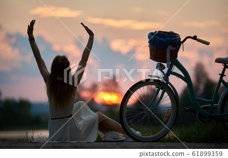 Slender girl sits with her back arms raised near retro bicycle with basket enjoying colorful sky with clouds at sunset Slender girl sits with her back arms raised near retro bicycle with basket enjoying colorful sky with clouds at sunset 61899359