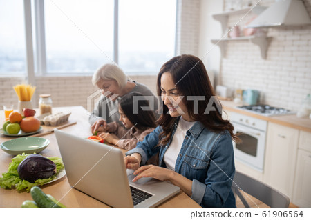 Mom at laptop in kitchen, grandmother with granddaughter slicing salad. Mom at laptop in kitchen, grandmother with granddaughter slicing salad. 61906564
