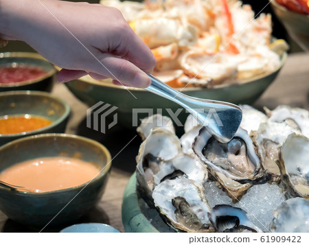 Hand of Asian woman pick a tasty fresh shucked oyster on ice with tongs. Hand of Asian woman pick a tasty fresh shucked oyster on ice with tongs. 61909422