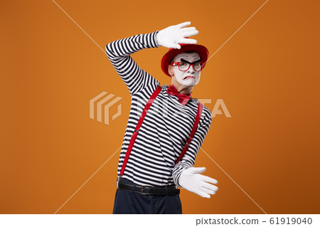 Mime man in vest and red hat with invisible glass in hands on orange background 61919040