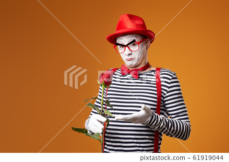 Serious mime in red hat and vest with rose in hands on orange background Serious mime in red hat and vest with rose in hands on orange background 61919044