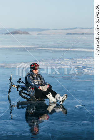 Woman sitting near her bike on ice. The girl cyclist stopped to rest. She sits on the wheel and enjoys the beautiful view of the sunset. Woman sitting near her bike on ice. The girl cyclist stopped to rest. She sits on the wheel and enjoys the beautiful view of the sunset. 61942856