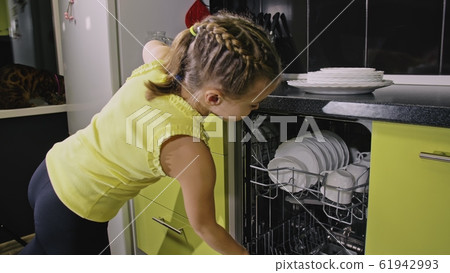 Smart girl learning to use dishwasher. Stylish modern Built In Kitchen Appliances in green black. Child is putting clean dishes. 61942993