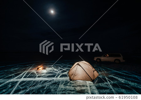 Fire on ice at night. Campground on ice. Tent stands next to bonfire. Lake Baikal. Nearby there is car. Shelter tent and ice are illuminated from the inside. Beautiful bonfire on real cracked ice. 61950108