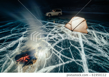 Fire on ice at night. Campground on ice. Tent stands next to bonfire. Lake Baikal. Nearby there is car. Shelter tent and ice are illuminated from the inside. Beautiful bonfire on real cracked ice. Fire on ice at night. Campground on ice. Tent stands next to bonfire. Lake Baikal. Nearby there is car. Shelter tent and ice are illuminated from the inside. Beautiful bonfire on real cracked ice. 61950112