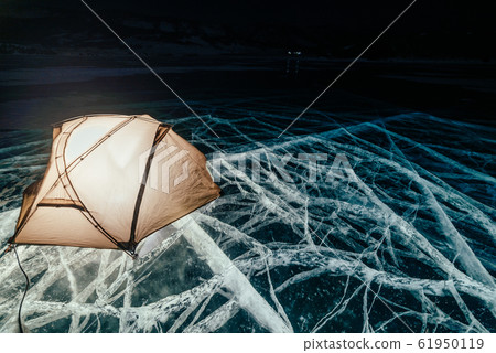 Fire on ice at night. Campground on ice. Tent stands next to bonfire. Lake Baikal. Nearby there is car. Shelter tent and ice are illuminated from the inside. Beautiful bonfire on real cracked ice. 61950119