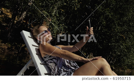 Woman shooting on handheld film gimbal stabilization for smartphone. Girl lie sunbed on pier make selfie. Blogger broadcast video vlogging. Woman shooting on handheld film gimbal stabilization for smartphone. Girl lie sunbed on pier make selfie. Blogger broadcast video vlogging. 61950715