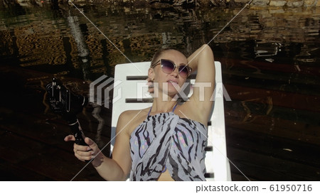 Woman shooting on handheld film gimbal stabilization for smartphone. Girl lie sunbed on pier make selfie. Blogger broadcast video vlogging. Woman shooting on handheld film gimbal stabilization for smartphone. Girl lie sunbed on pier make selfie. Blogger broadcast video vlogging. 61950716