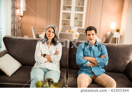 Mature couple sitting on the sofa after quarrel and looking at camera Mature couple sitting on the sofa after quarrel and looking at camera 61955304