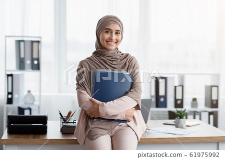 Smiling Arabic Businesswoman Holding Folders And Posing Near Workplace In Office Smiling Arabic Businesswoman Holding Folders And Posing Near Workplace In Office 61975992